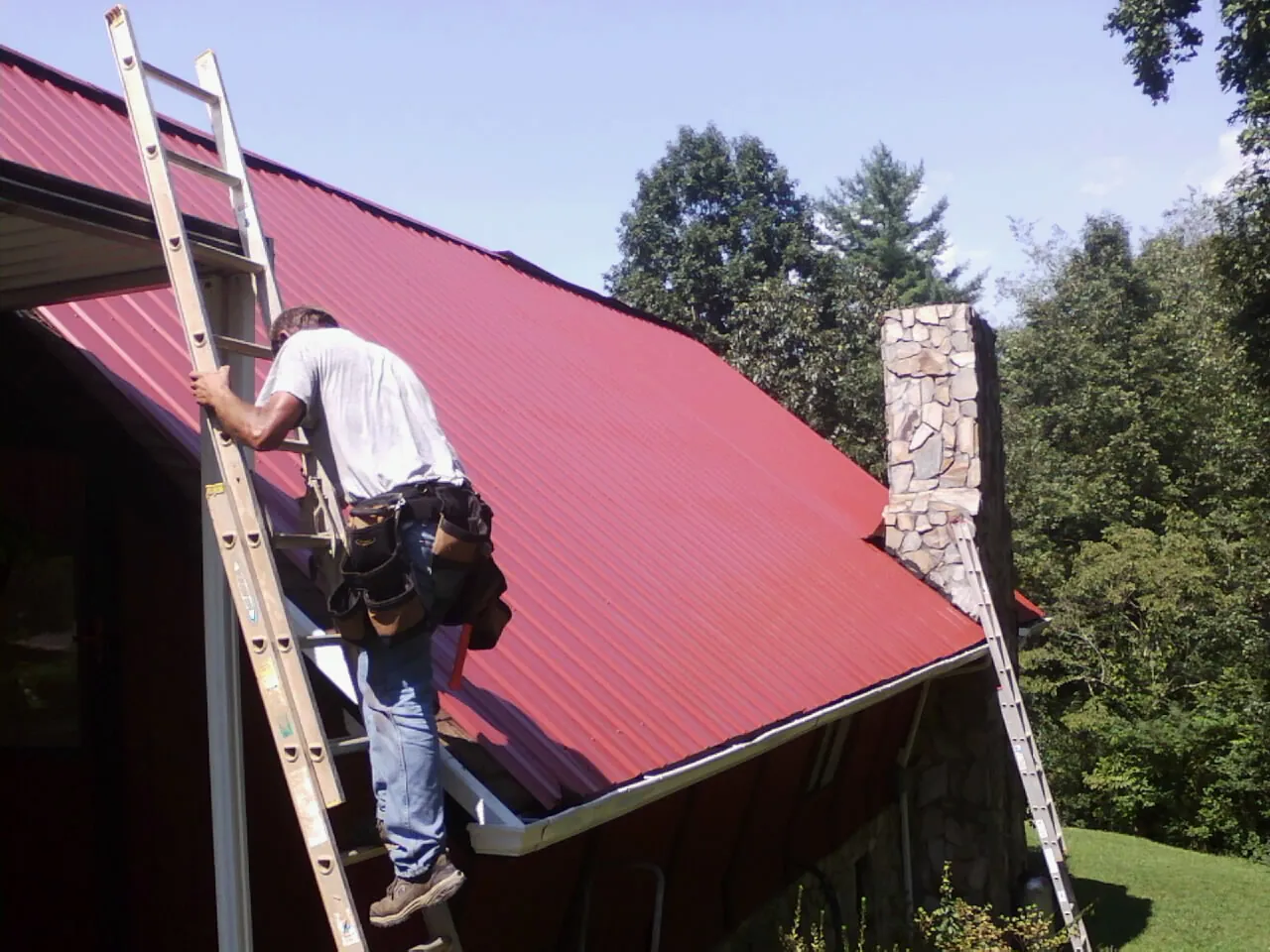 Roofer on a ladder beside a red metal roof and stone chimney — Soffit & Fascia Repair by Roofing Brothers in Keller